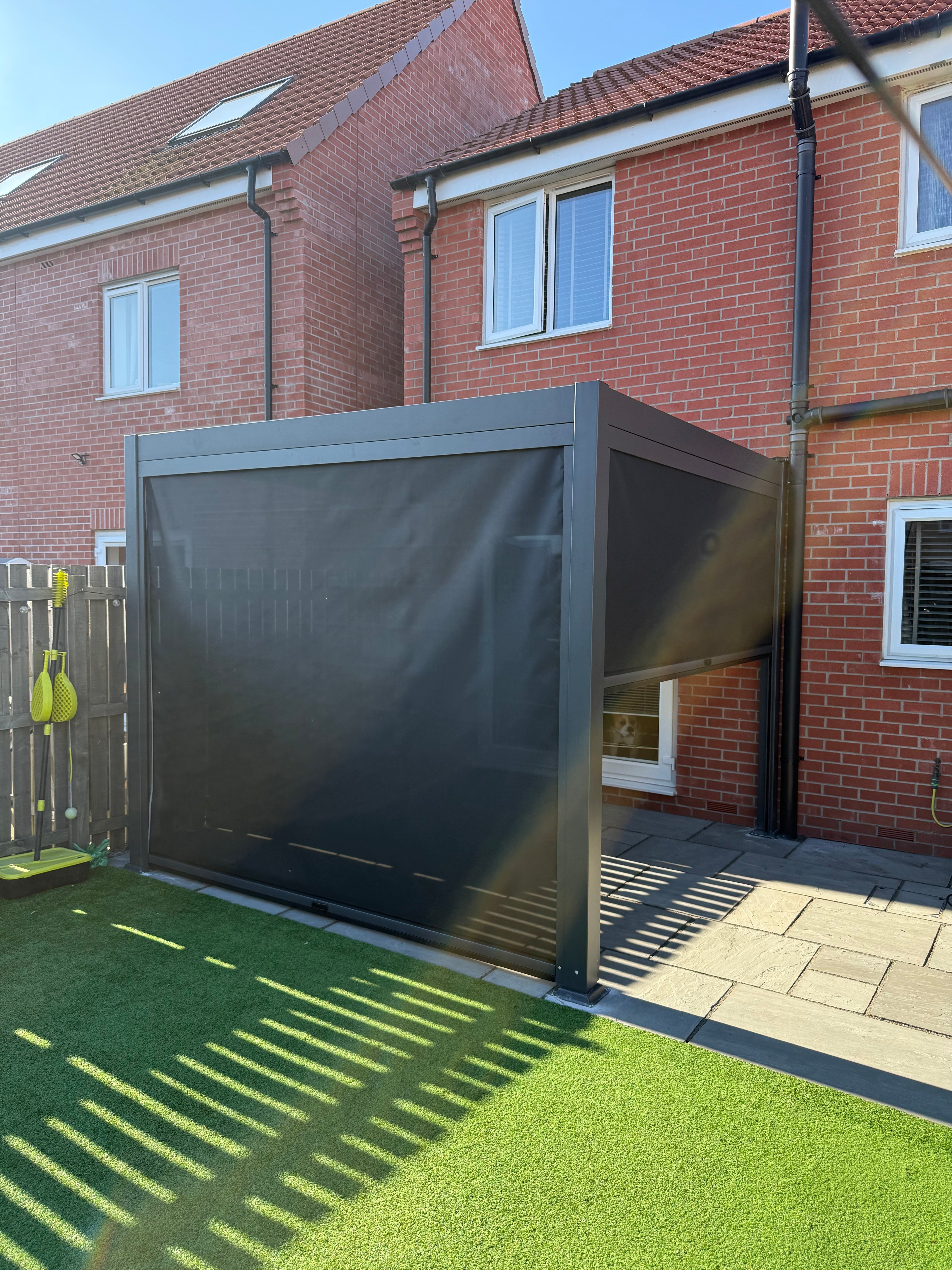 Anthracite grey aluminium louvred pergola installed against a red-brick house in Hull, over a paved patio beside artificial lawn