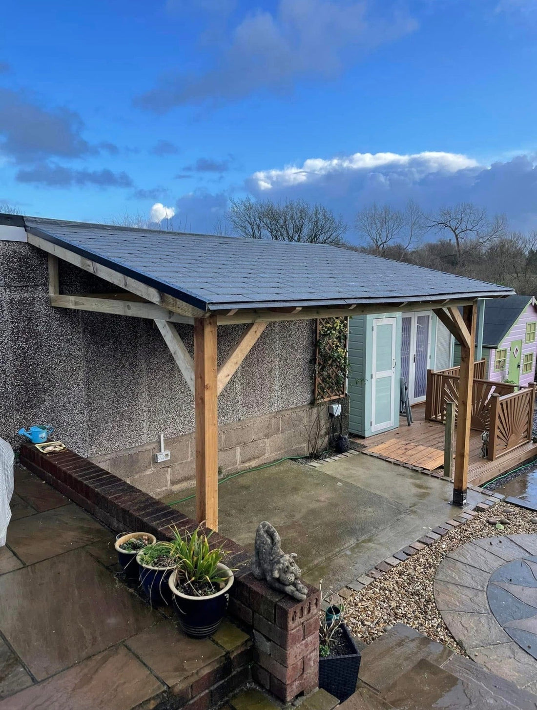 Bespoke lean-to timber pergola built on site in Preston Lancashire — heavy timber posts with diagonal knee bracing, slate-effect shingle roof and canal views beyond the garden